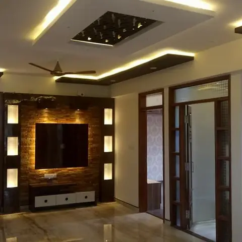 Living room with a multi-tier cove-lit false ceiling with a jaali cutout centerpiece, a stone-clad TV wall with lit display niches, and marble flooring.