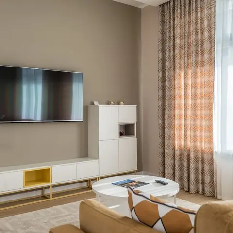 Living room with a mustard-gold TV console and taupe wall, patterned geometric curtains layered over sheers by the window.