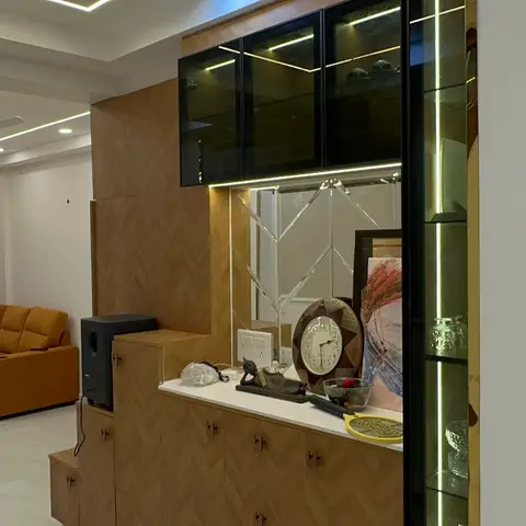 Living room with a mustard sofa and nested gold-frame coffee tables, seen past a herringbone-panelled console topped with a backlit glass display cabinet.