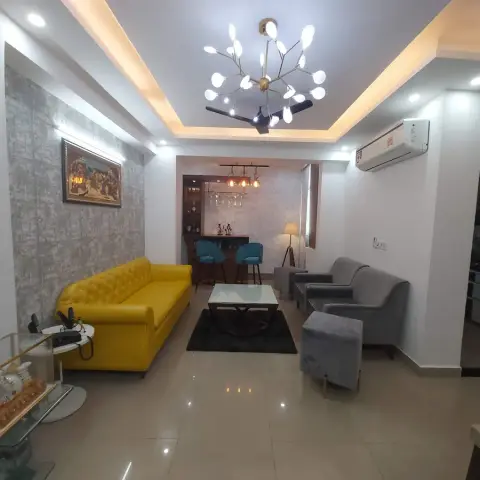 Living room with a mustard-yellow tufted sofa and grey armchairs around a black rug, cove-lit ceiling with a branched pendant chandelier
