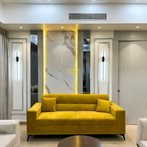Living room with a mustard-yellow sofa set facing a backlit marble feature wall flanked by mirror panels and slim wall sconces, fluted panelling by the door