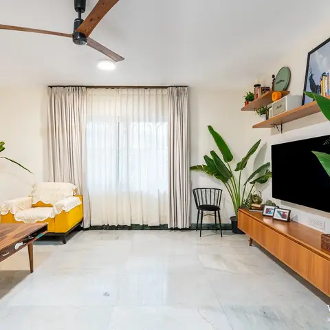 Living room with a mustard-yellow sofa and armchair under crochet throws, a mid-century wood TV console styled with books and plants