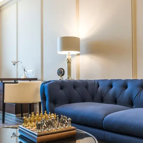 Living room with a navy blue tufted chesterfield sofa, panel-moulded walls, a drum-shade table lamp and a chess set styled on the glass coffee table