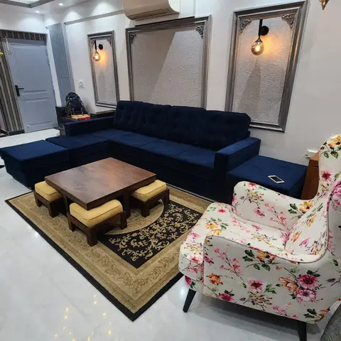 Living room with a navy velvet tufted sectional, a floral wingback armchair, and a low wooden coffee table with four stools on a patterned rug.