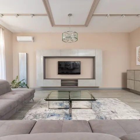 Living room with a peach-toned coffered ceiling, a stone-look TV feature wall, a glass coffee table on a blue abstract rug, and grey sectional sofas.