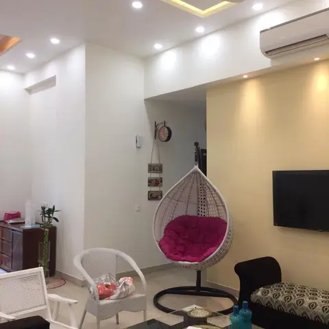Living room with a rattan hanging egg chair with a fuchsia cushion, a mustard accent wall behind the wall-mounted TV, and a black glass coffee table.