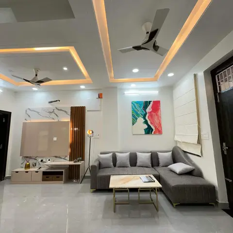 Living room with a stepped cove-lit ceiling and two fans, a marble-and-wood TV feature wall, and a grey L-shaped sofa facing a gold-framed coffee table.