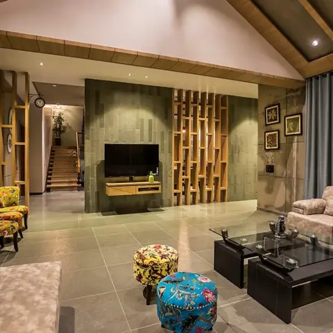 Living room with a stone-clad TV wall flanked by a wood slat screen, sloped wood ceiling, and colorful floral ottomans in front of a beige sofa.