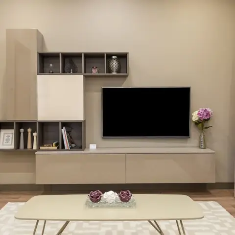 Living room with a taupe asymmetric wall-mounted TV unit featuring open display cubbies, a mauve patterned curtain, and an oval coffee table on a cream rug.