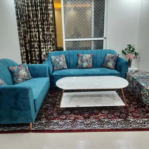 Living room with a teal channel-tufted sofa set, jewel-toned floral cushions and a matching ottoman, a marble-top coffee table, and a red patterned rug.