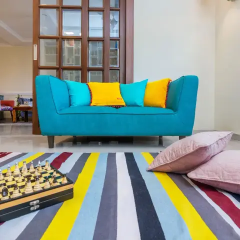 Living room with a teal sofa styled with yellow and turquoise cushions, a striped multicolor dhurrie rug with a chess-set vignette, pink floor cushions