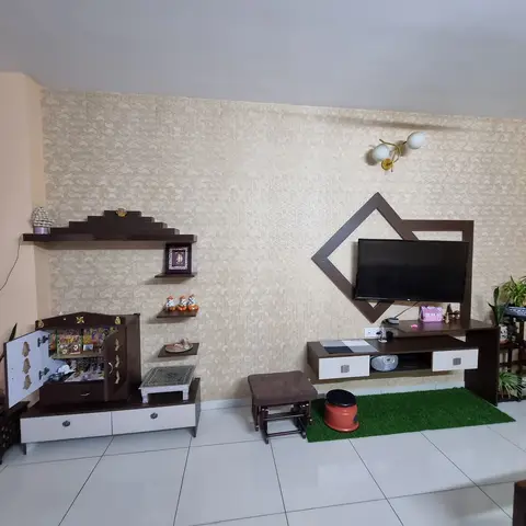 Living room with a textured geometric wallpaper feature wall, a diamond-framed wood TV console on one side and a dark-wood pooja cabinet with tiered display