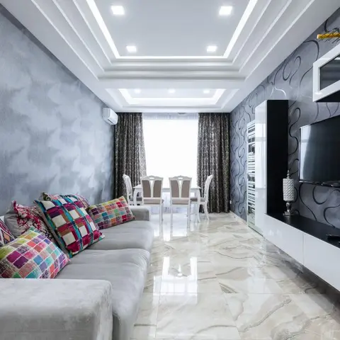Living room with a textured grey accent wall, colourful patterned throw pillows and a glossy black-and-white TV console against swirl-pattern wallpaper.