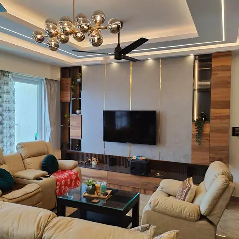 Living room with a walnut-and-brass TV feature wall, cove-lit tray ceiling, a bubble chandelier, and beige reclining sofas around a glass-top coffee table.
