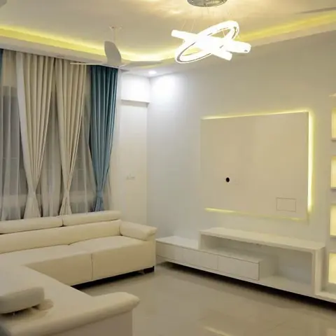 Living room with a white L-shaped sofa, teal-and-cream layered curtains and a backlit TV wall unit with glowing display niches.