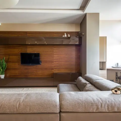 Living room with a wood-panelled TV wall featuring a backlit floating display shelf, a low glossy console, and a grey sectional sofa in the foreground.