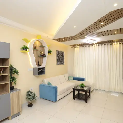 Living room with a wood-slat angled false ceiling, a cream-and-blue L-shaped sofa and a wall-mounted yellow-accented pooja niche with a Buddha statue.