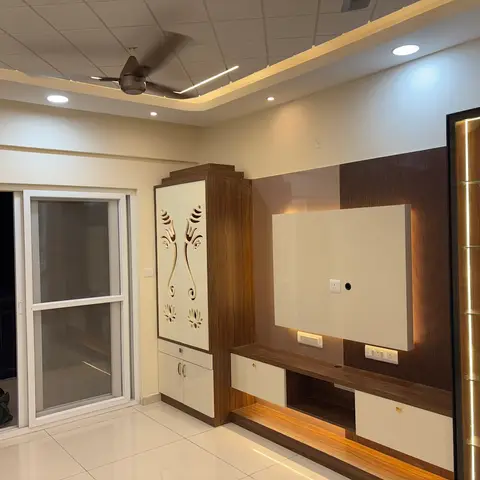 Living room with a wood-veneer TV wall, still wrapped in protective film, beside a tall white pooja cabinet with carved lotus motifs and a backlit glass