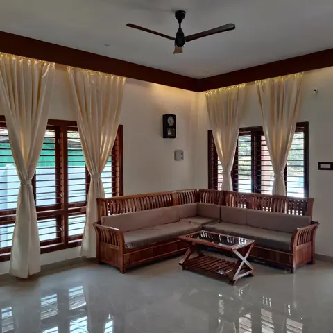 Living room with a wooden slatted L-shaped sofa, cream curtains framing louvred wood-shutter windows, and a glass-and-wood coffee table on a polished floor.