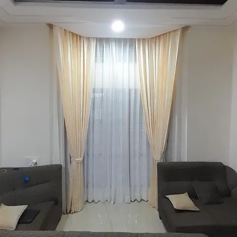 Living room with an angled wood-slat ceiling detail around a ceiling fan, floor-length cream curtains framing a tall window, and grey L-shaped sofa seating.