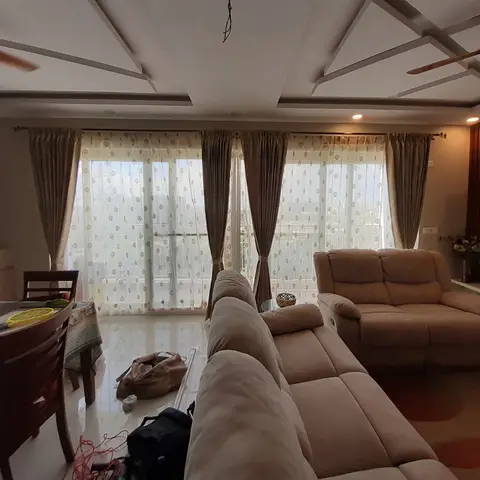Living room with an angular geometric false ceiling and floor-length patterned sheer curtains framing a city-view balcony, a beige reclining sofa set in front
