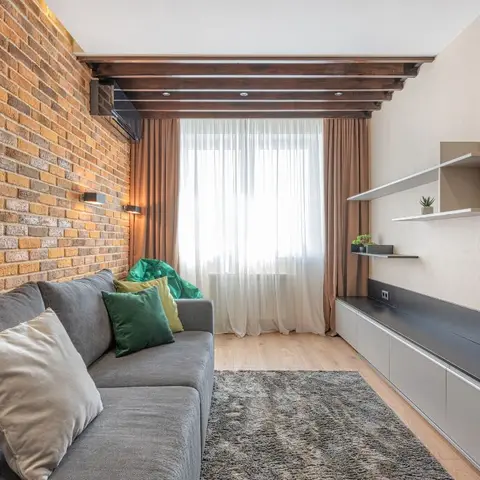 Living room with an exposed brick accent wall, dark wood beam ceiling, a grey sectional sofa with green and mustard cushions, and a floating black TV console.