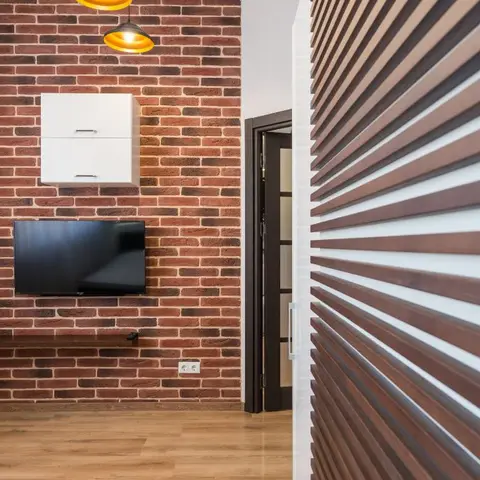 Living room with an exposed red-brick feature wall, wall-mounted TV and white cabinet, amber pendant lights, and a fluted wood partition wall in the foreground.