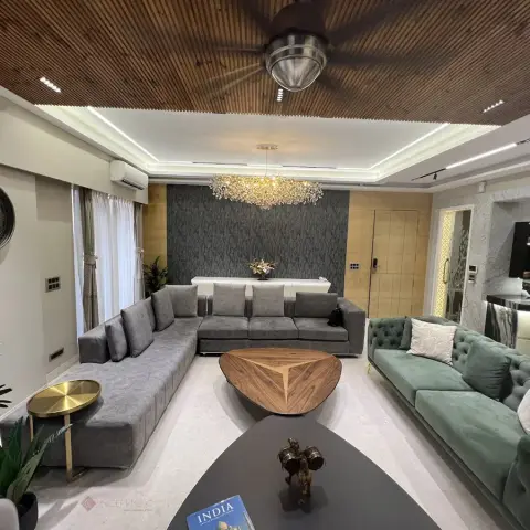 Living room with an L-shaped grey sofa facing a tufted sage-green sofa, a triangular wood coffee table, a crystal chandelier under a wood-slat ceiling
