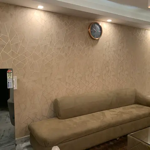 Living room with an olive-green suede sofa against a beige geometric crackle-pattern wallpaper wall inlaid with gold lines, a round brass-rimmed wall clock