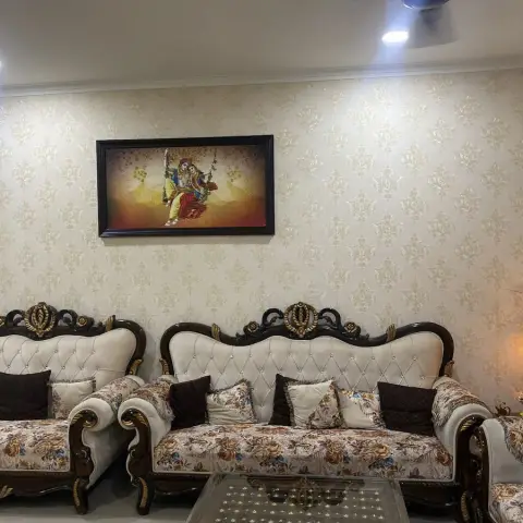 Living room with an ornate carved-wood sofa set in floral upholstery beneath a framed Radha-Krishna artwork on damask wallpaper.