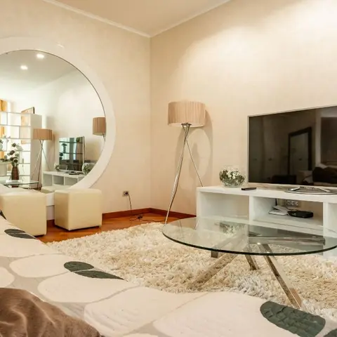 Living room with an oversized round wall mirror above two cream ottomans, a glass-top coffee table on a cream shag rug, and a white low TV console.