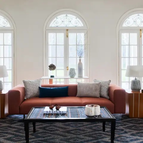 Living room with arched French windows, a terracotta three-seater sofa, dark patterned area rug, and a large ceramic floor planter