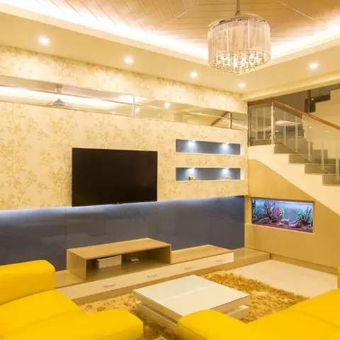 Living room with bright yellow leather sofas facing a mirrored TV wall with floral wallpaper, a built-in aquarium beneath the glass-railed staircase
