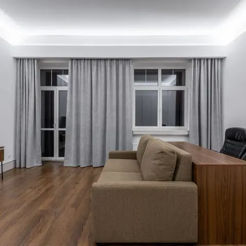 Living room with cove-lit ceiling, floor-to-ceiling grey curtains over twin windows, a wood TV console, and a wooden desk with an office chair behind the sofa.