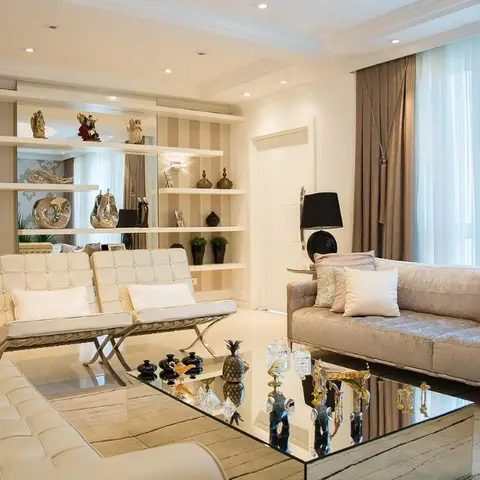 Living room with cream Barcelona-style chairs, a mirrored coffee table styled with decorative objects, and a floating shelf display wall.