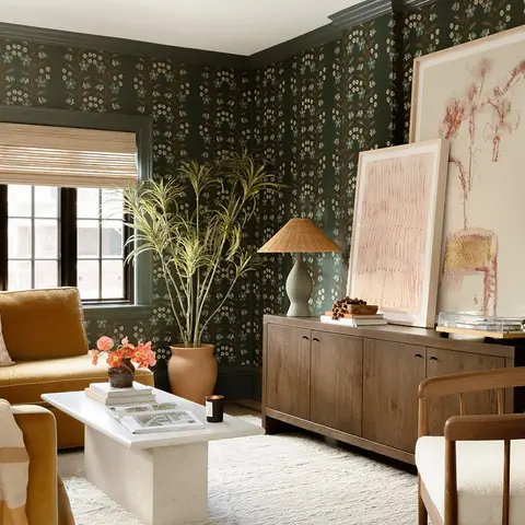 Living room with dark green botanical wallpaper, a mustard velvet sofa, a wood console styled with leaning framed art, and a travertine coffee table.