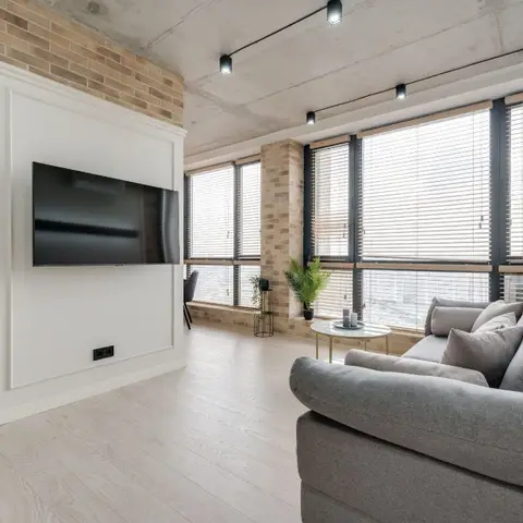 Living room with exposed concrete ceiling and track lighting, brick-clad columns, floor-to-ceiling windows with venetian blinds, and a grey fabric sofa.