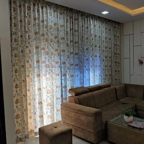 Living room with floral-print sheers drawn across an entire wall, a brown suede L-sofa with matching cube ottoman and a glass-top storage coffee table under a