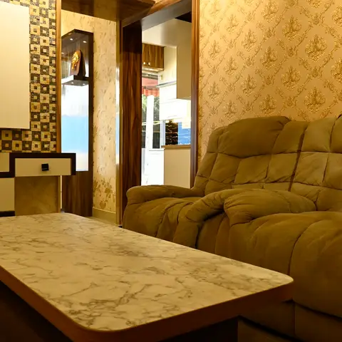 Living room with gold damask wallpaper, a tufted olive recliner sofa and a marble-top coffee table facing a checkered-tile TV console wall with a lit display