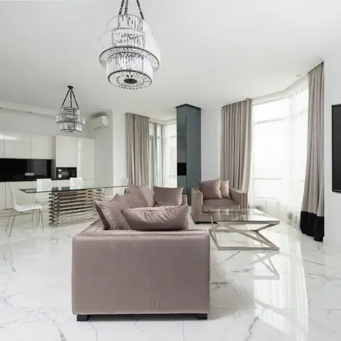 Living room with mauve velvet sofas and a geometric brass coffee table on glossy white marble flooring, tiered crystal chandelier above, kitchen visible behind