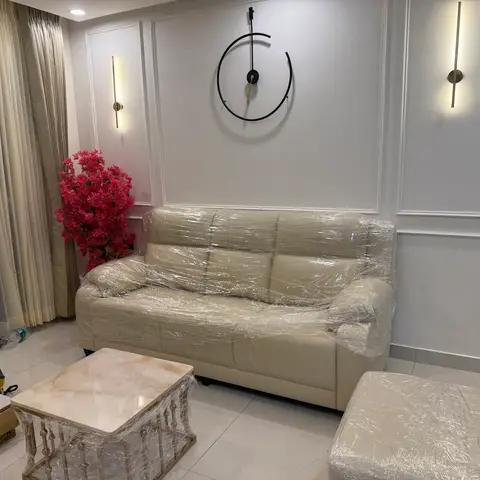 Living room with panel-moulded walls, twin linear brass wall sconces, and a beige leather sofa still wrapped for delivery beside a marble-and-brass coffee
