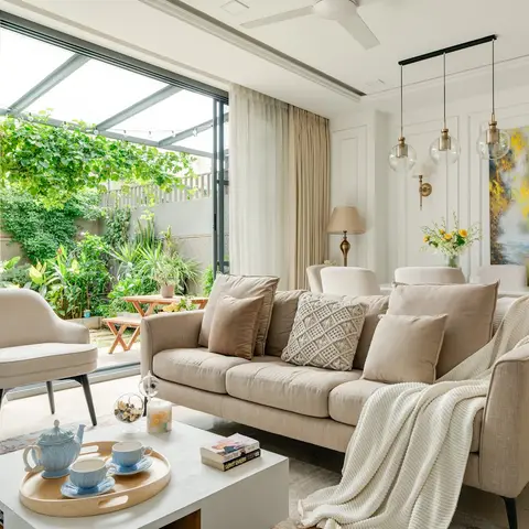 Living room with panelled wainscot walls, a beige sofa layered with textured cushions and a knit throw, opening through glass doors to a green garden terrace.
