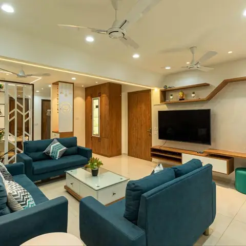 Living room with three teal sofas around a white glass coffee table, an angled floating wood TV shelf, and a white geometric jaali room divider.