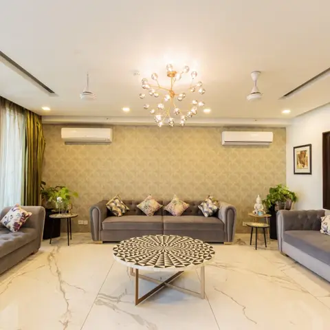 Living room with three tufted grey velvet sofas around a round mosaic-top coffee table, patterned gold-and-olive wallpaper wall, a gold laser-cut jaali screen
