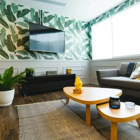 Living room with tropical leaf-print wallpaper above white wainscoting, a wall-mounted TV, and a grey sofa with mustard-yellow accent cushions on a striped rug.