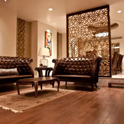 Living room with tufted brown leather chesterfield sofas and a carved wooden coffee table, divided from the dining area by a gold geometric jaali screen