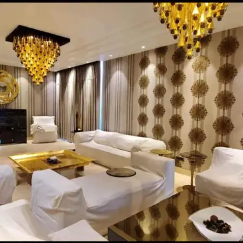 Living room with twin gold cluster chandeliers, gold medallion-pattern wallpaper, and white slipcovered sofas around a mirrored gold coffee table.