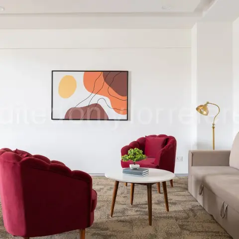 Living room with two maroon tufted velvet armchairs and a matching sofa around a round wood-legged coffee table, beneath an abstract orange-toned art print