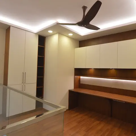 Loft study nook with a long built-in wooden desk under lit overhead cabinets, beside a glass-railed staircase landing.