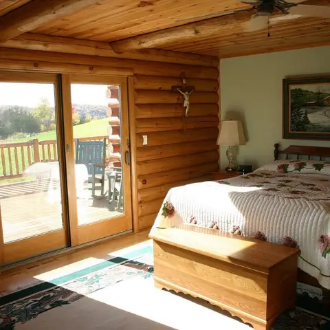 Log-cabin bedroom with a spindle-back wooden bed under a floral quilt, a cedar blanket chest at its foot and sliding glass doors onto a deck over open country.